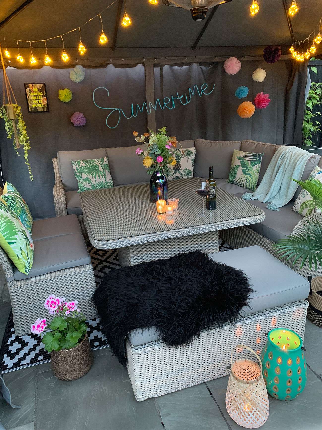 Patio table and chairs under a fabric gazebo decorated with colour pompoms, festoon lights, plants, tropical cushions and lanterns with red wine and candles on the table.