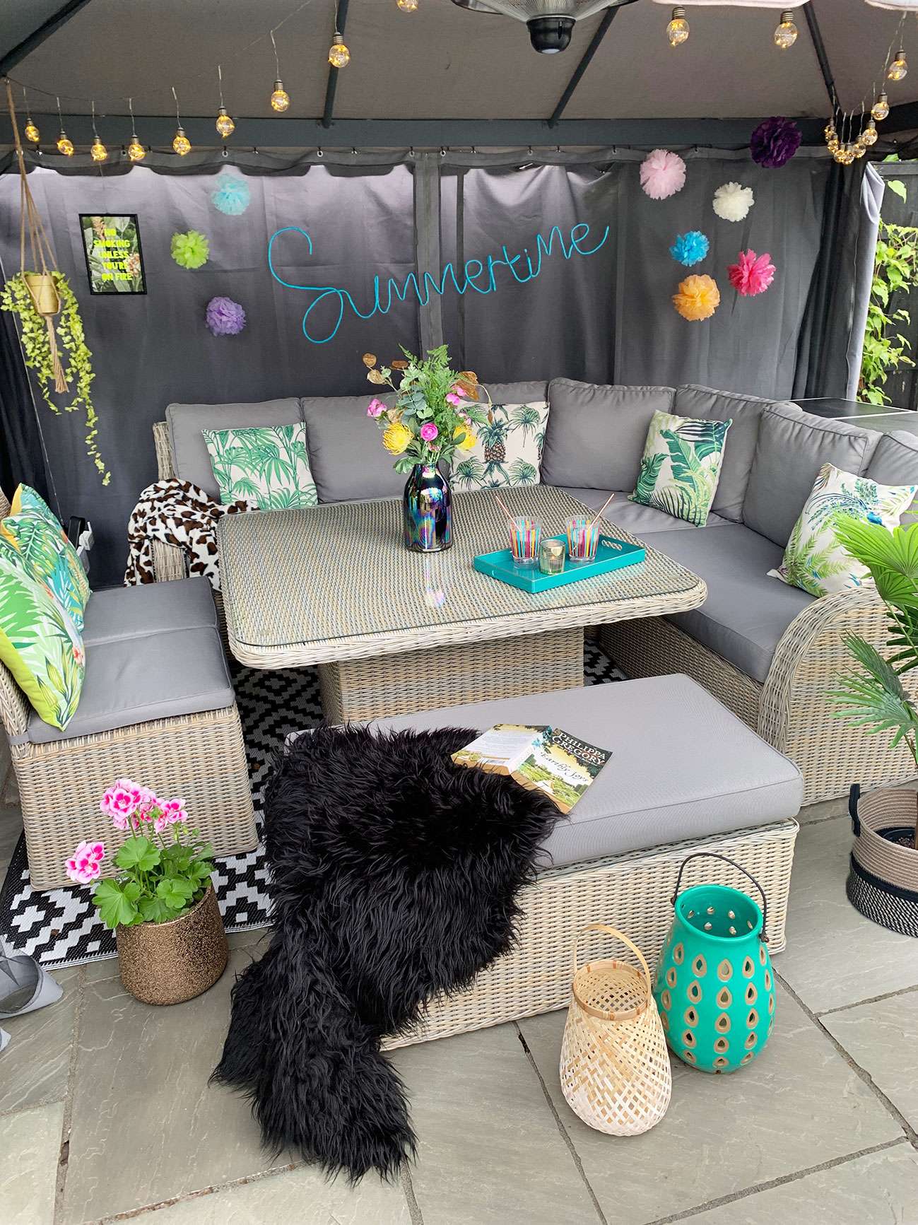 Patio table and chairs under a fabric gazebo decorated with colour pompoms, festoon lights, plants, tropical cushions and lanterns with drinks on the table.