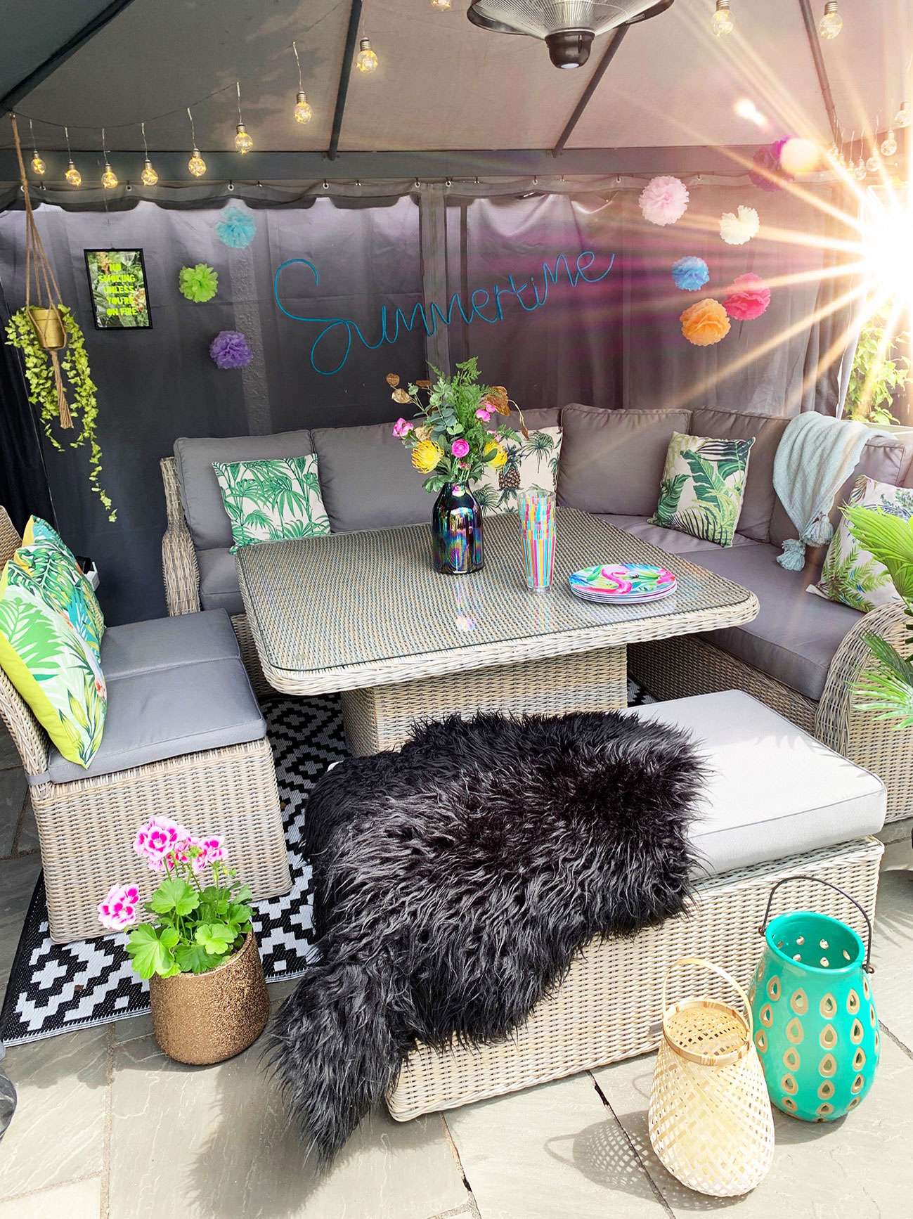 Patio table and chairs under a fabric gazebo decorated with colour pompoms, festoon lights, plants, tropical cushions and lanterns with flamingo plates and cups on the table.