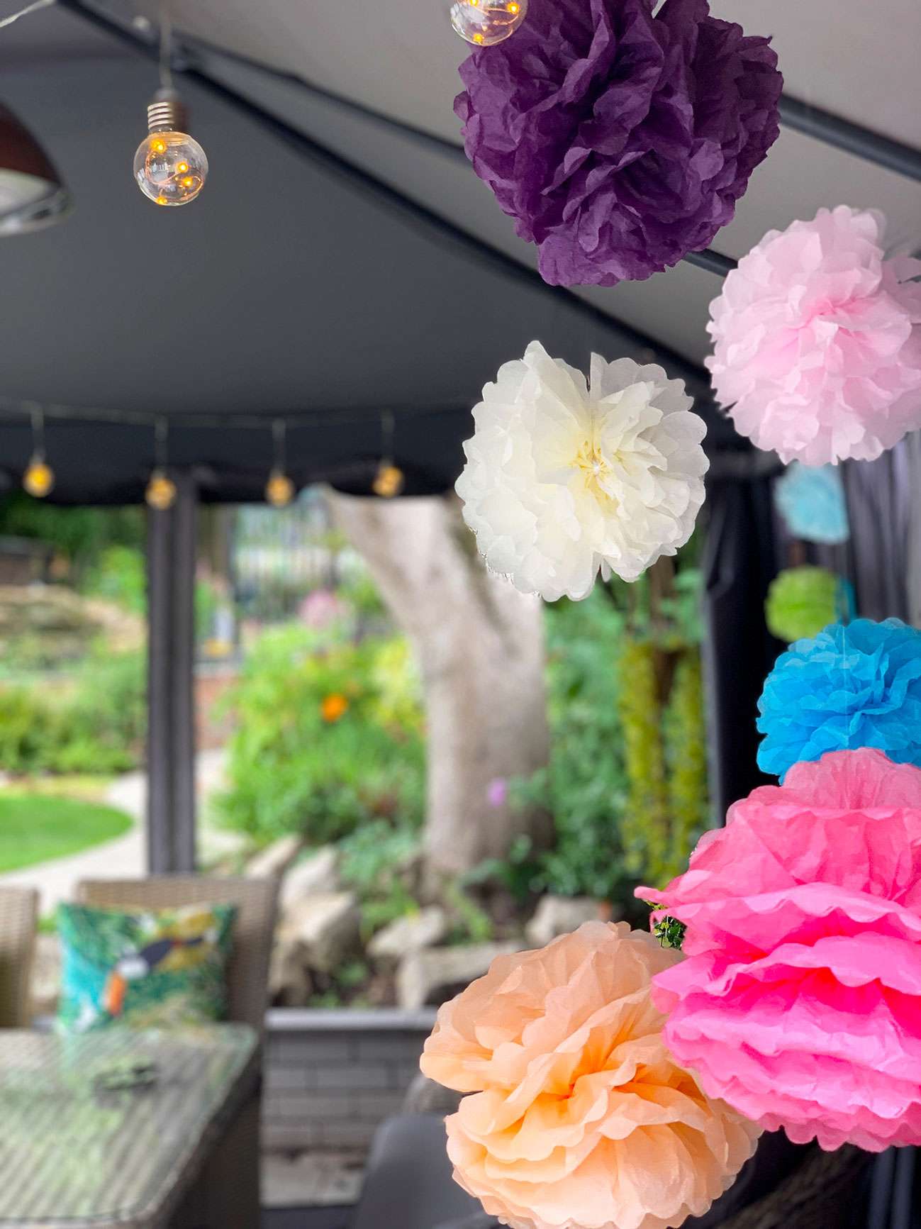 Colour paper pompoms in a gazebo with festoon lighting and patio set in background.