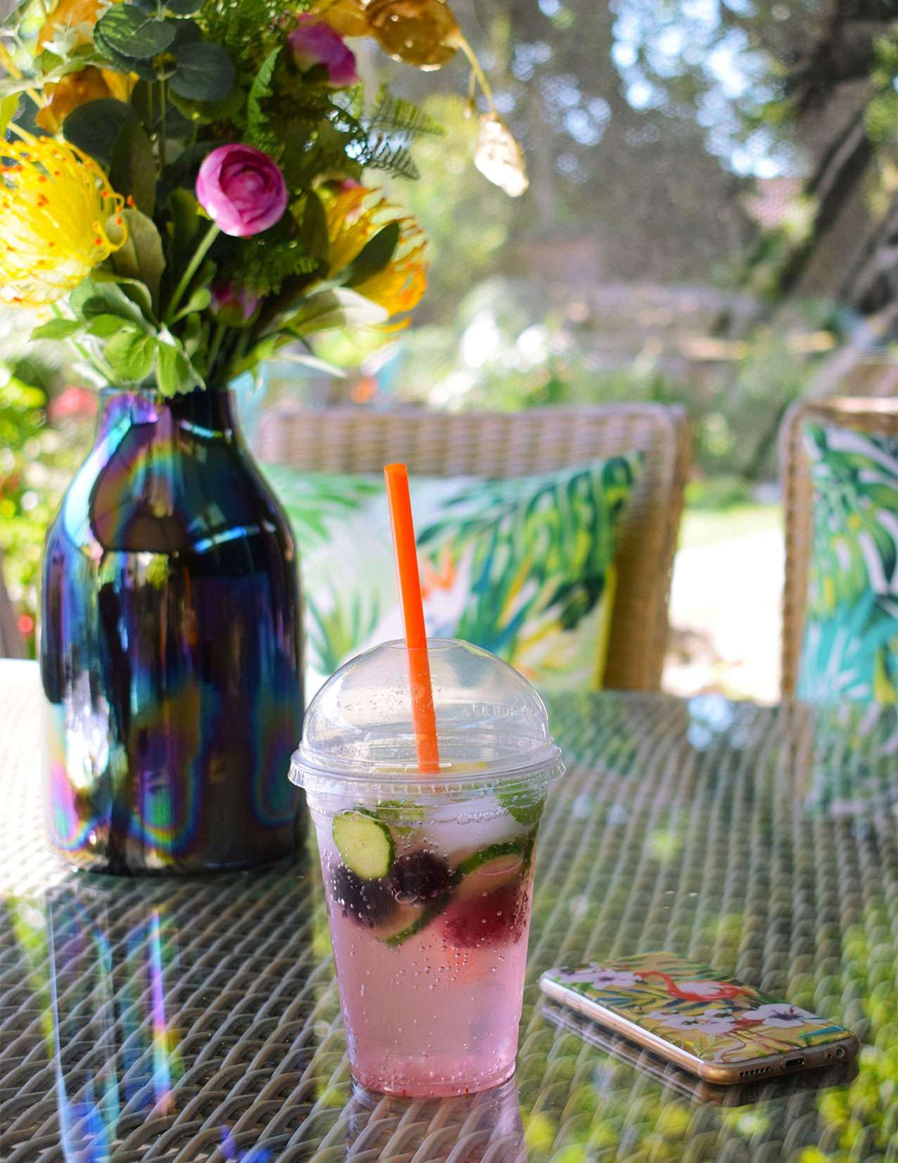Artificial flowers, pink soda water, and phone on garden table with tropical cushions.