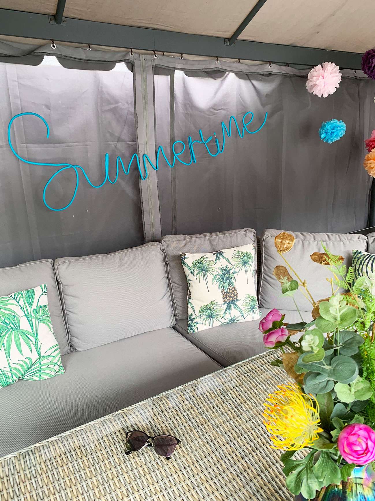 Garden patio set with flowers and sunglasses on the table, tropical cushions, and DIY summertime outdoor decorative sign.
