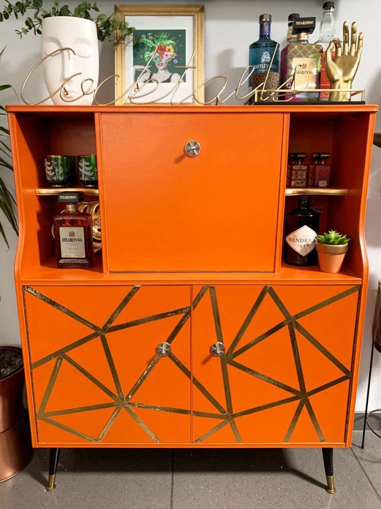 Upcycled vintage G Plan cocktail cabinet in burnt orange with gold geometric pattern with alcohol bottles on shelves.