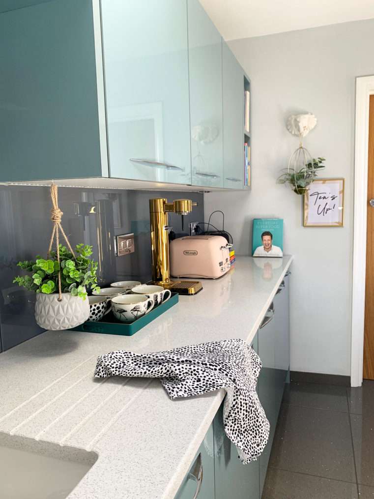 corner of kitchen worktop with pink toaster, coffee mugs on teal tray, herb planter and animal print tea towel.