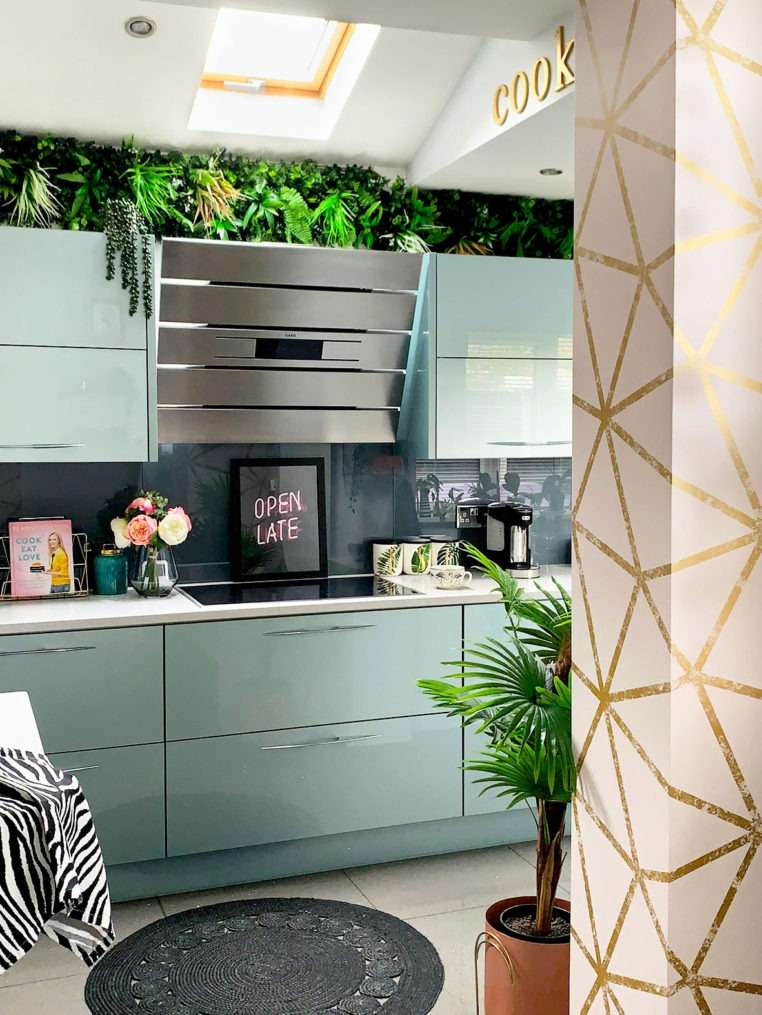 part of kitchen with hob, extraction, and blue fitted kitchen with living wall above wall cupboards and pink and bold geometric wallpaper on kitchen column.