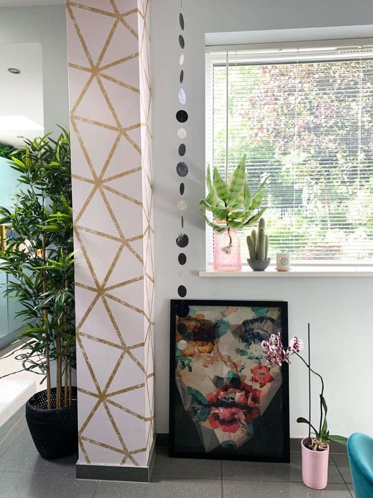 room column with blush and gold geometric wallpaper with lady face picture next to it leaned against a wall with window above.
