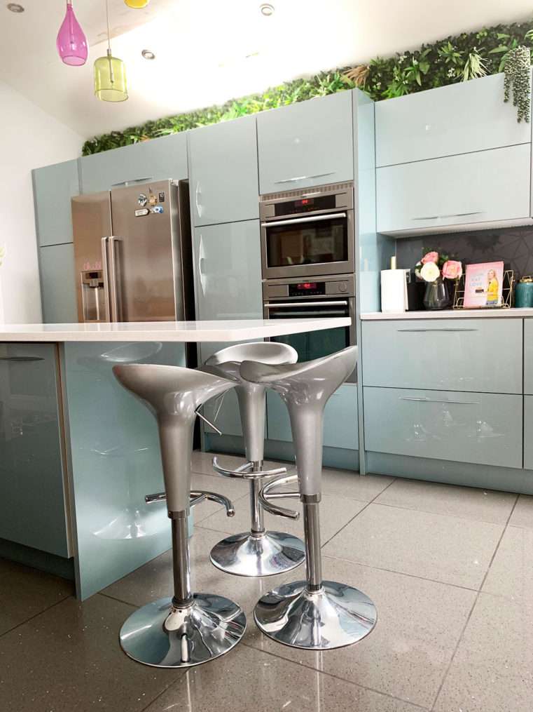 kitchen island with silver stools below white quartz counter top, on grey quartz floor tiles with kitchen units and oven and fridge behind.
