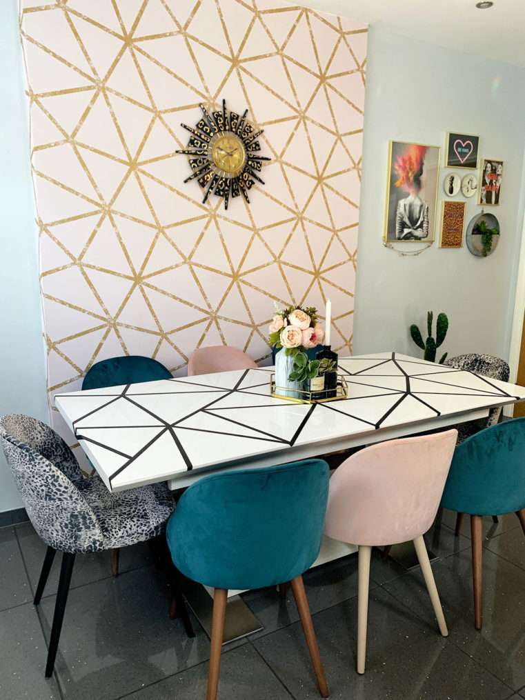 dining area with pink and gold feature wallpaper behind table with geometric pattern and velvet dining chairs in blush, teal, and leopard print.