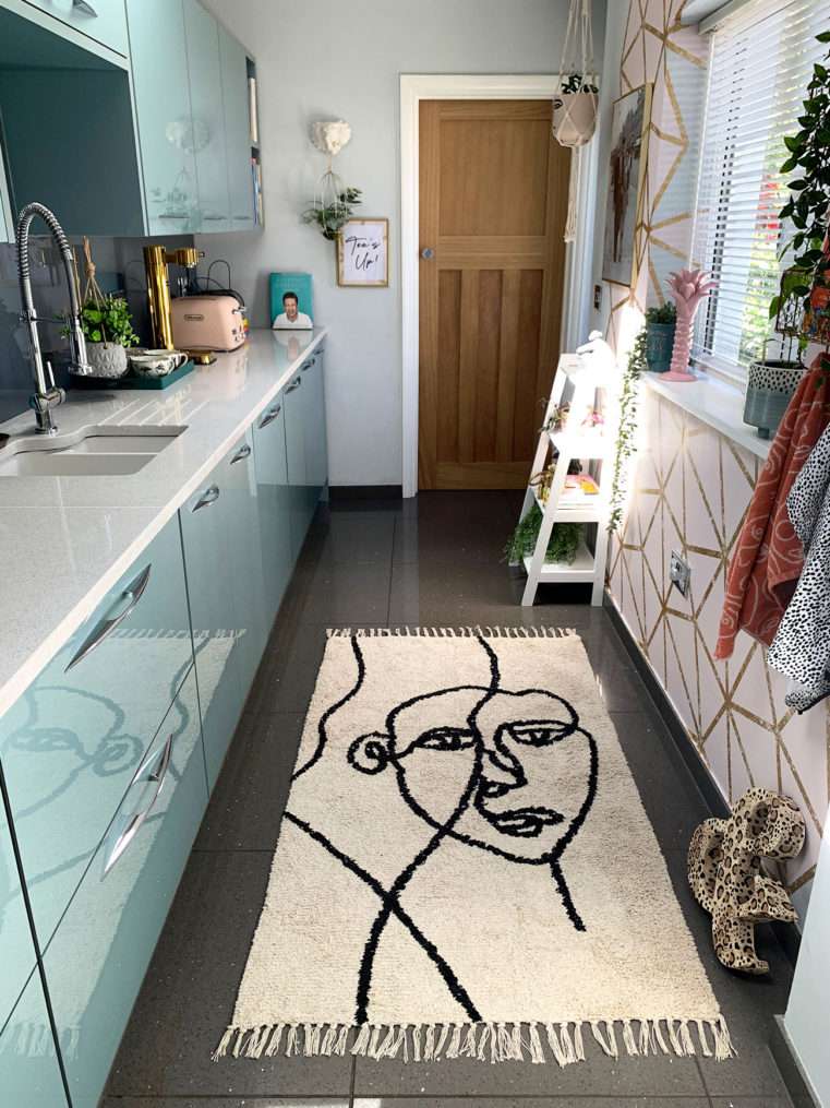 end of kitchen with blue cupboards and quartz worktop. Abstract face rug on grey quartz floor with pink and gold geometric wallpaper to the right around a window with accessories.