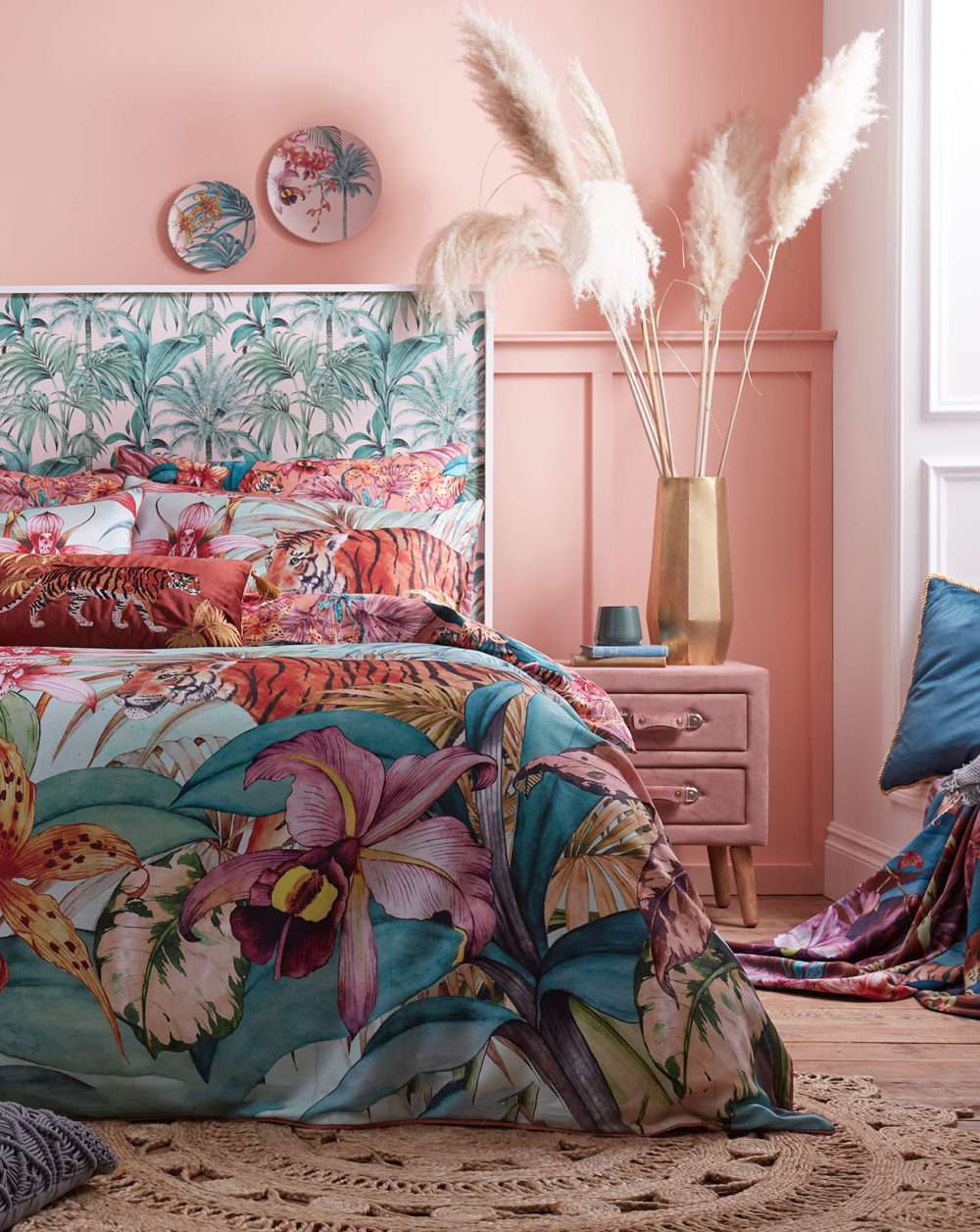 Corner of a pink bedroom with a wooden floor covered with a Jute rug, underneath a bed dressed with a tropical duvet set and a pink velvet side table topped with a gold vase filled with pampas grass.