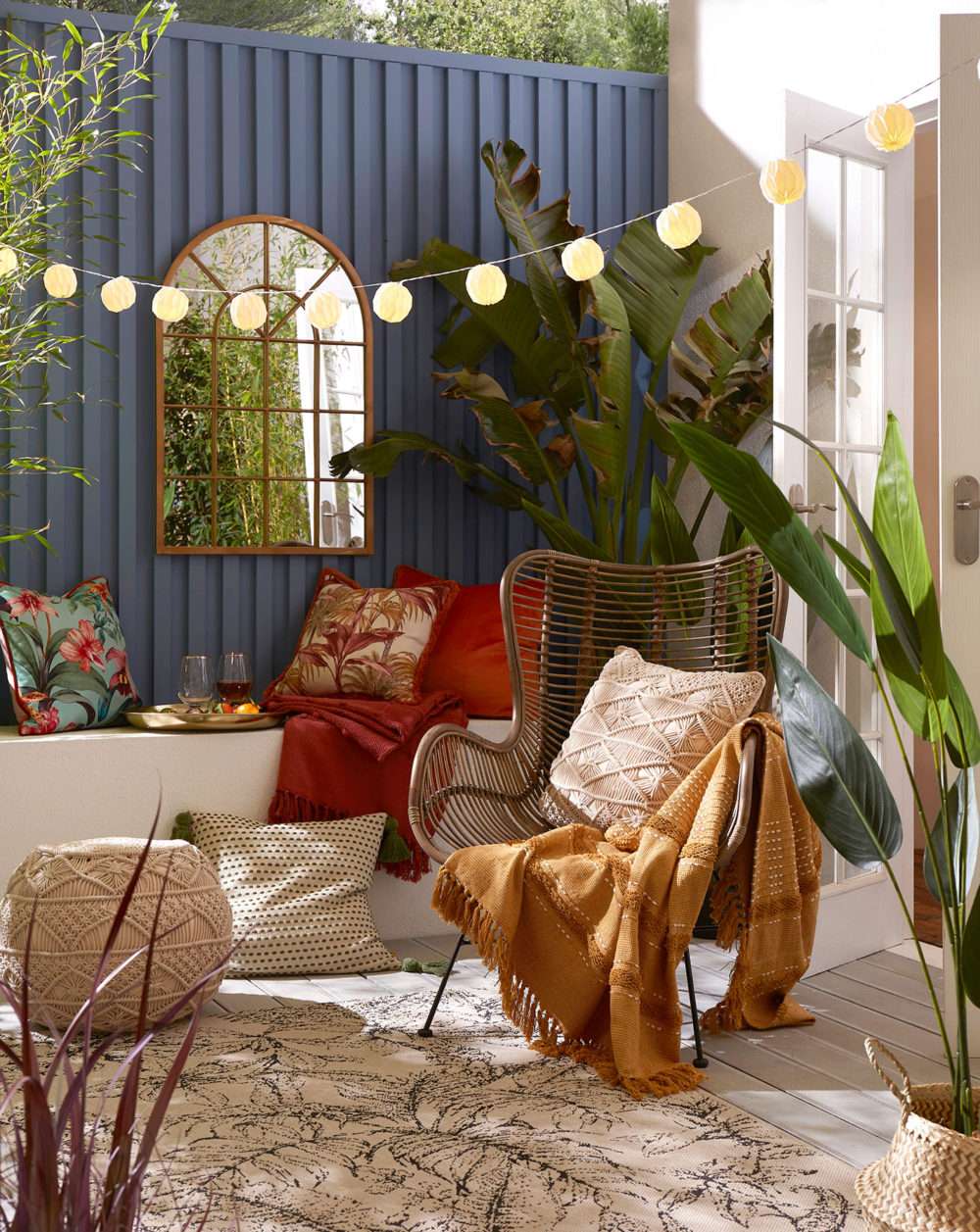 Garden patio with tropical cushions, bird of paradise plant and a rattan chair with an arch mirror and paper fairy lights.