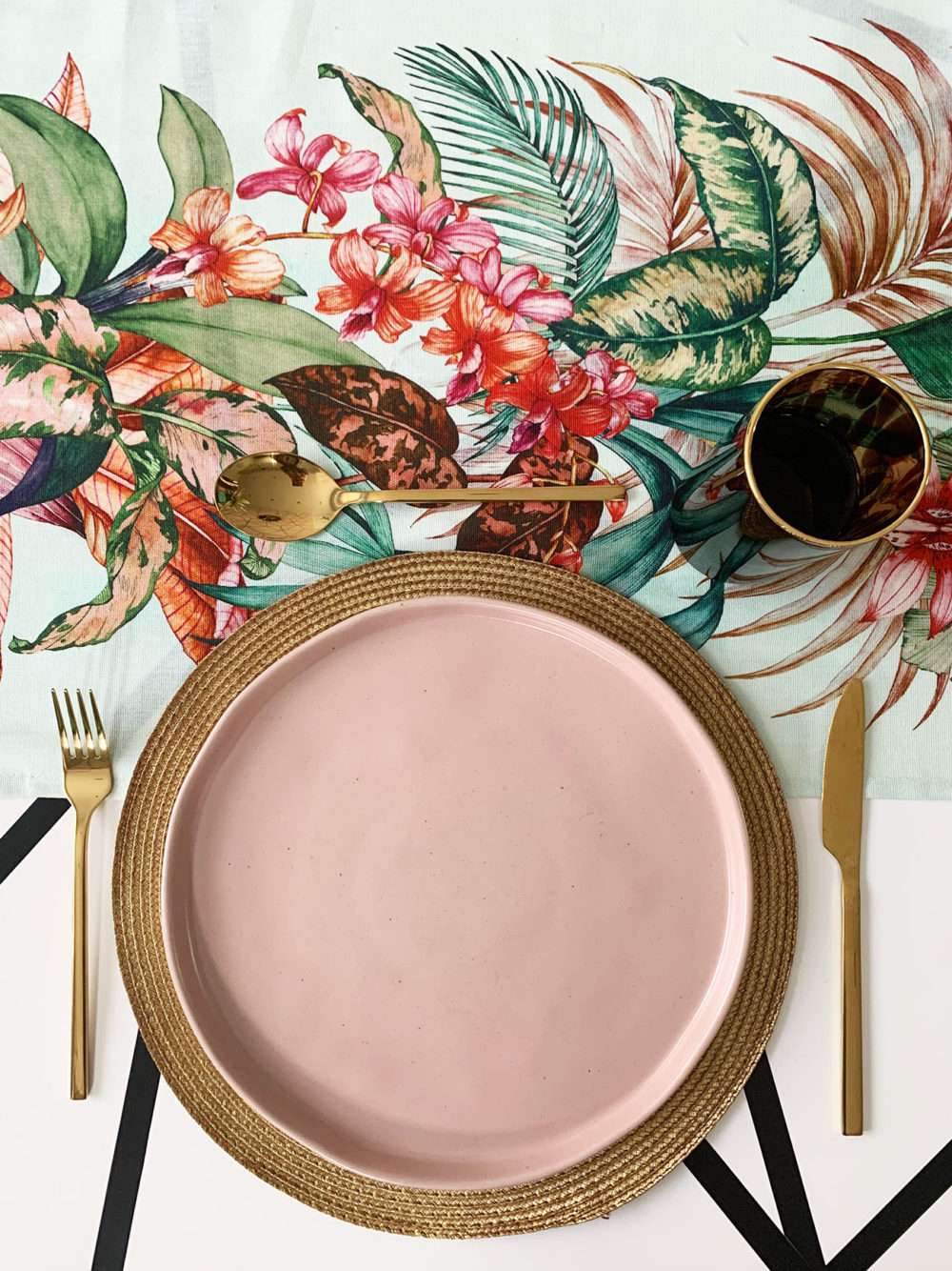 flatlay of dining table with a tropical table runner, blush plate on a gold placemat with gold cutlery and a tortoiseshell glass with a gold rim.