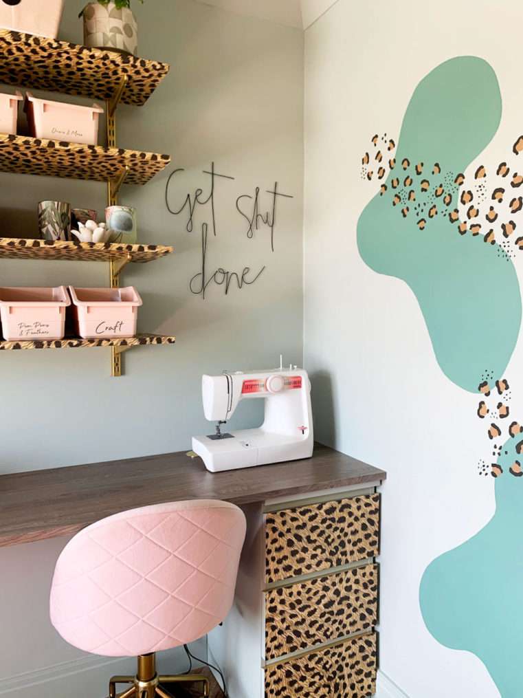 Corner of desk in soft green, blush, and leopard print decorated craft room with shelves, sewing machine, and black wire wall words saying 'Get Shit Done'.