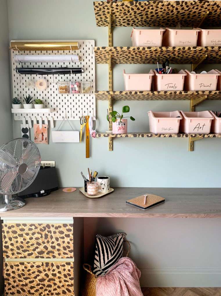 Corner of craft room with pin board, shelves, and worktop with belly basket containing cushion and blanket underneath.
