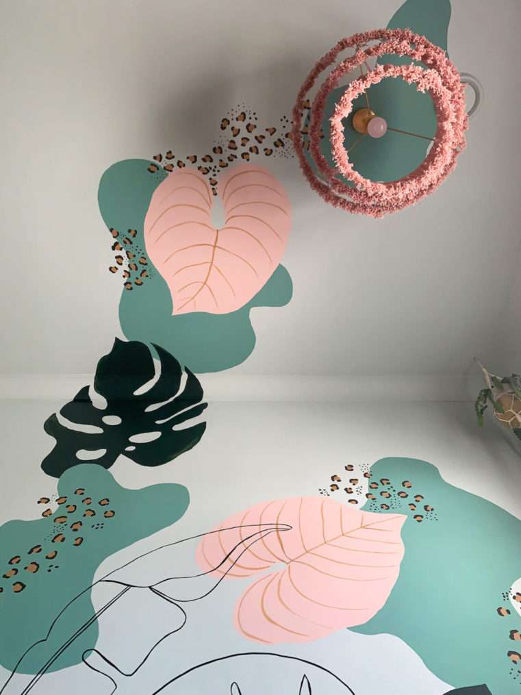Ceiling shot of handprinted tropical mural in green blush and leopard print next to dusty pink tassel lampshade.