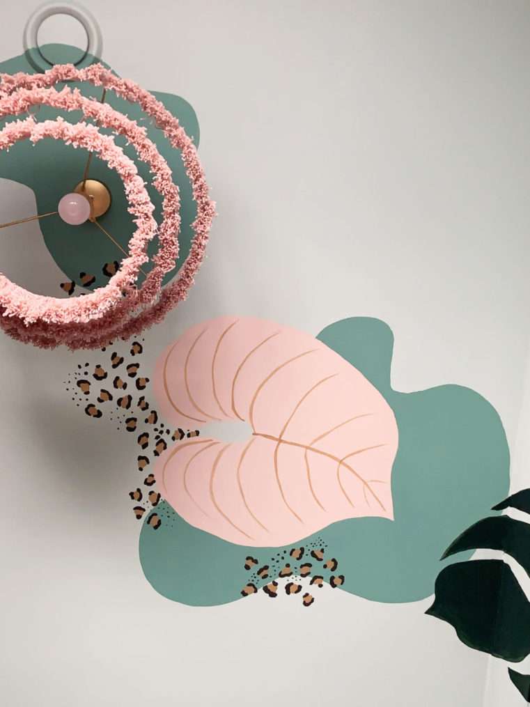 Close up ceiling shot of handprinted tropical mural in green blush and leopard print next to dusty pink tassel lampshade.