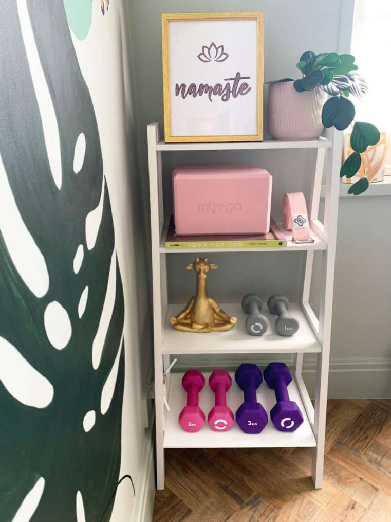 White ladder shelf with 'Namaste' text print, blush pant pot and yoga blocks, gold yoga giraffe ornament and dumbbells.