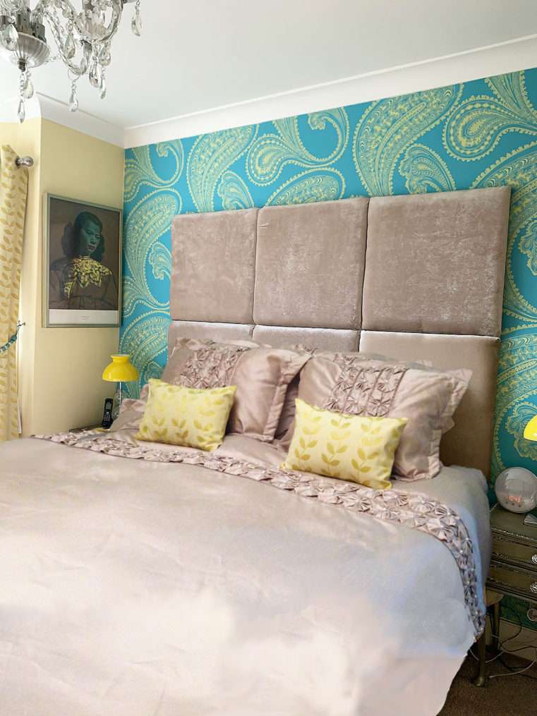 Bed with beige duvet cover and beige velvet headboard with turquoise and yellow feature wallpaper and 'The Chinese Girl' print.