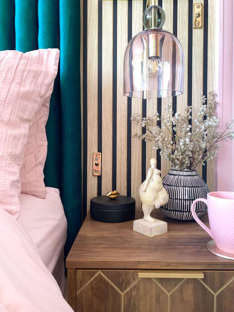 Close up of bedside table with blush glass wall light, lady figurine ornament, pink mug, monochrome vase containing dried mimosa stems, with a wood slat wall background.