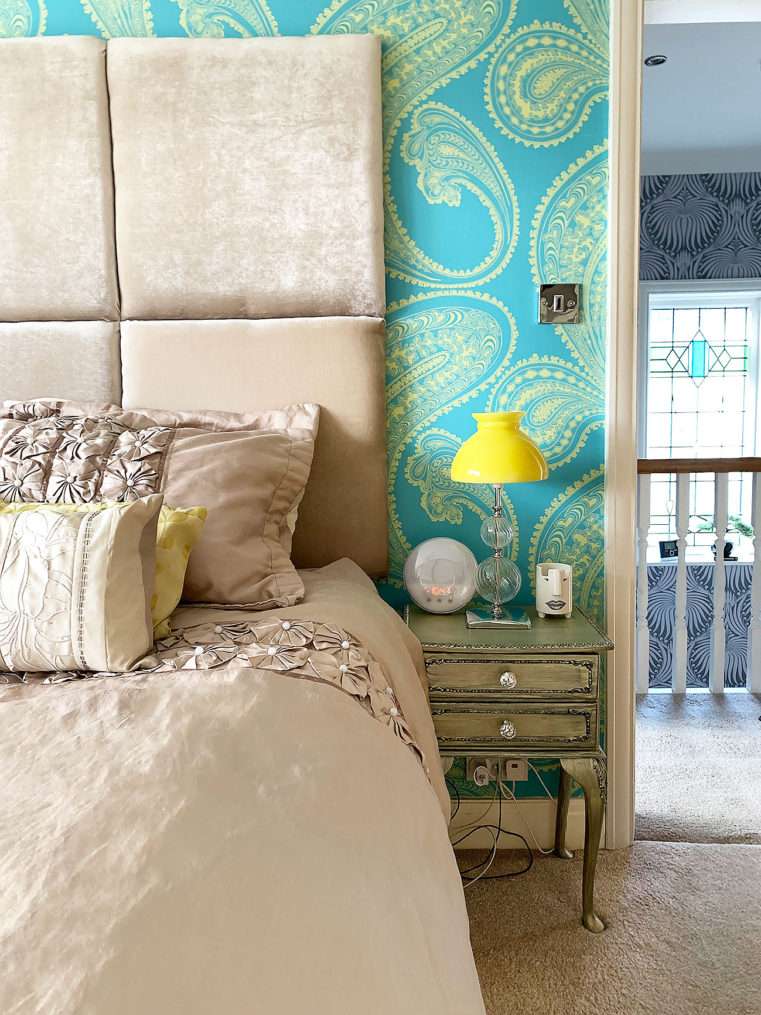 Close up of bedside with beige duvet cover and beige velvet headboard with turquoise and yellow feature wallpaper and yellow glass retro lampshade.