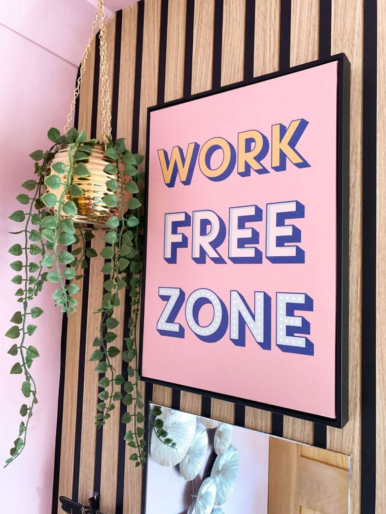 Close up of bedroom corner with gold hanging planter, typography pink 'Work Free Zone' print on wood slat wall.