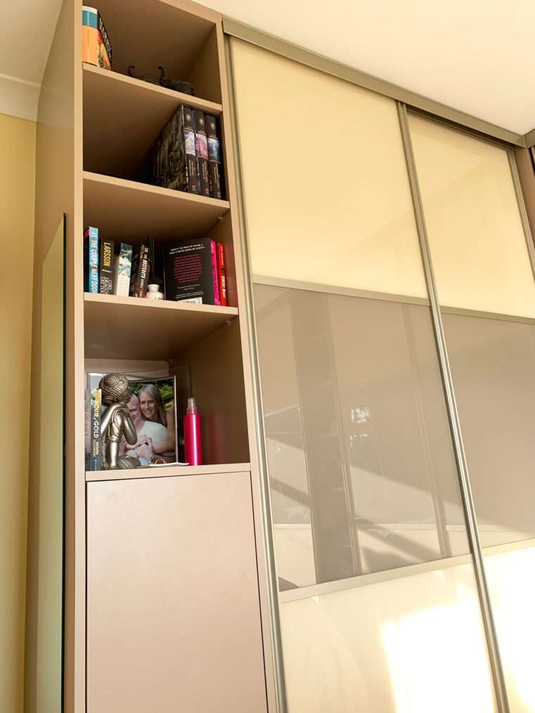 Beige glass sliding wardrobes with a left hand shelf with books on.