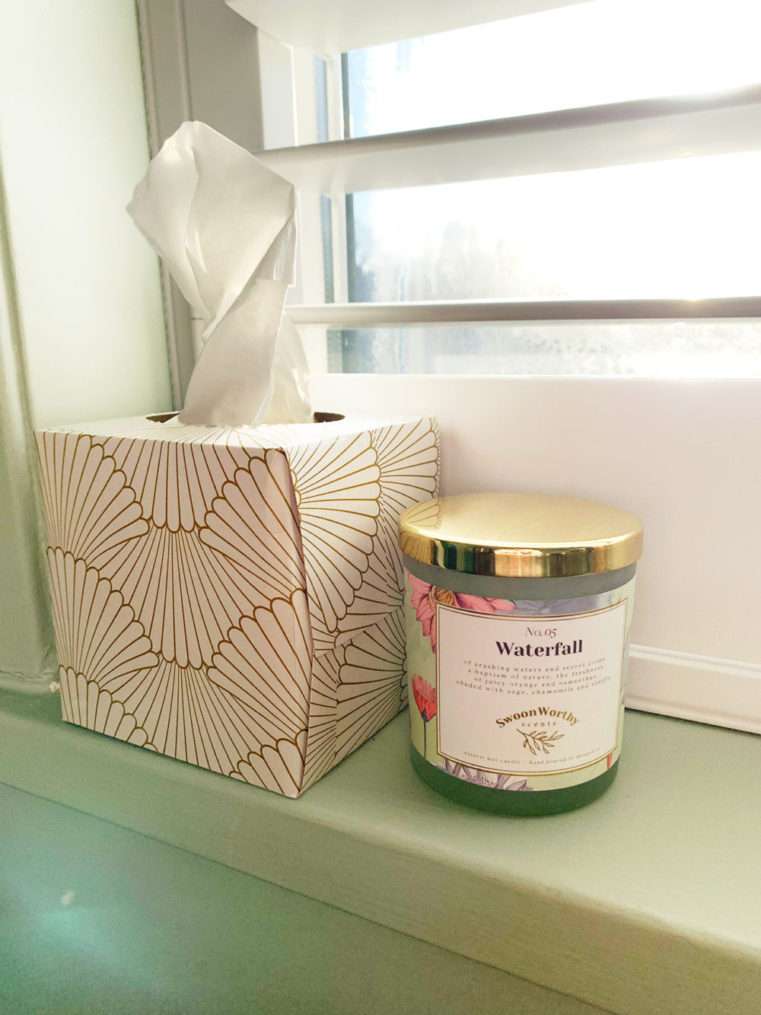 Close up of box of tissues and luxury candle on soft green windowsill.