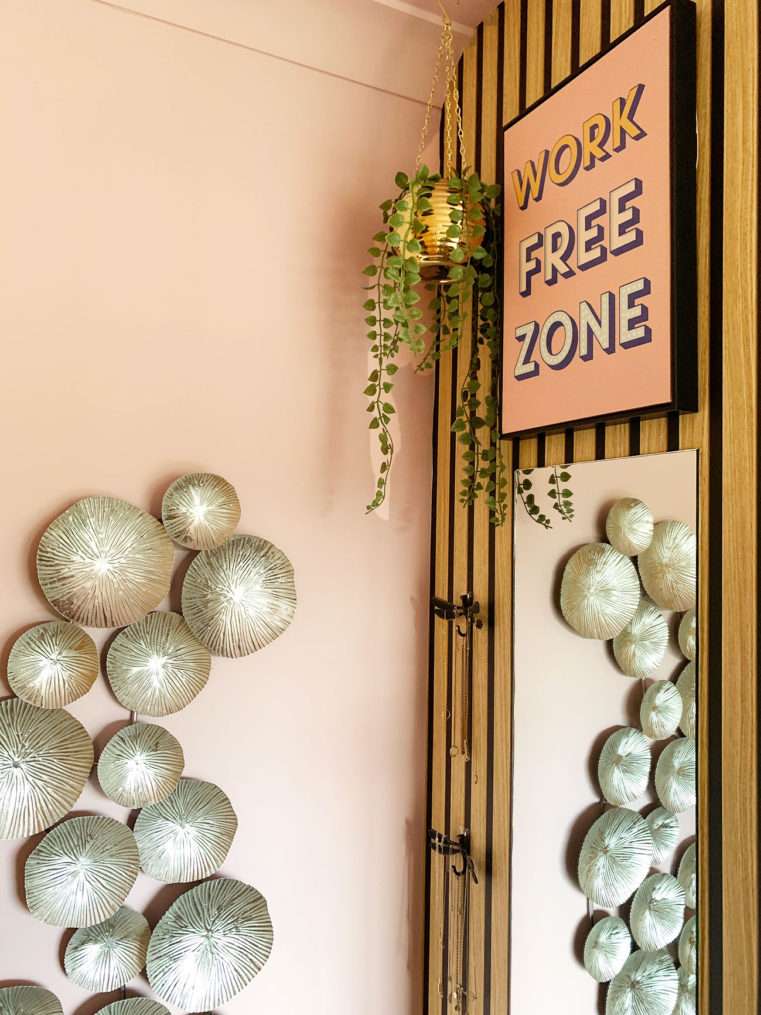 Close up of bedroom corner with Work Free Zone print on wood slat wall above full length mirror next to silver wall art on pink wall.