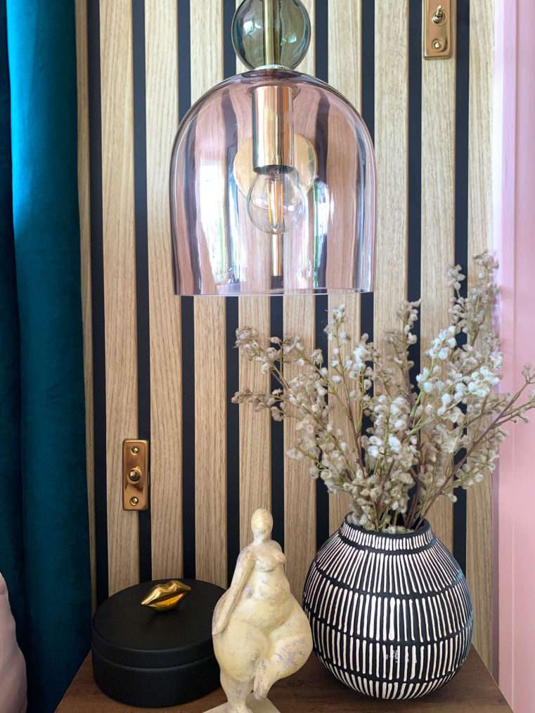 Boutique style bedside with pink and teal decor, blush glass and gold wall light, lady sculpture ornament, monochrome vase, dried mimosa stems, and slat wood wall.