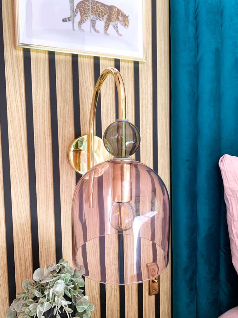 Close up of a bedside glass, blush and gold wall light with a slat wood panel wall.